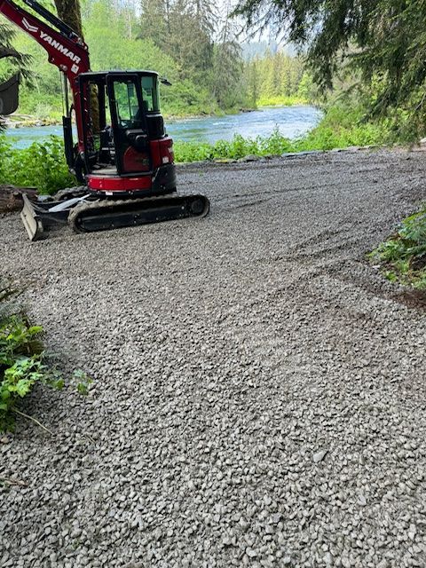 A red Yanmar mini excavator parked on a gravel area next to a flowing river surrounded by trees.