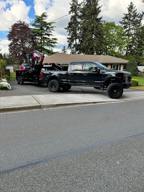 A black pickup truck parked on a residential street, towing a flatbed trailer loaded with a piece of heavy machinery.
