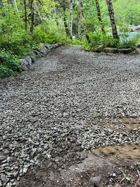 A gravel driveway under construction, bordered by large rocks, leading toward trees and a pond in a wooded area.