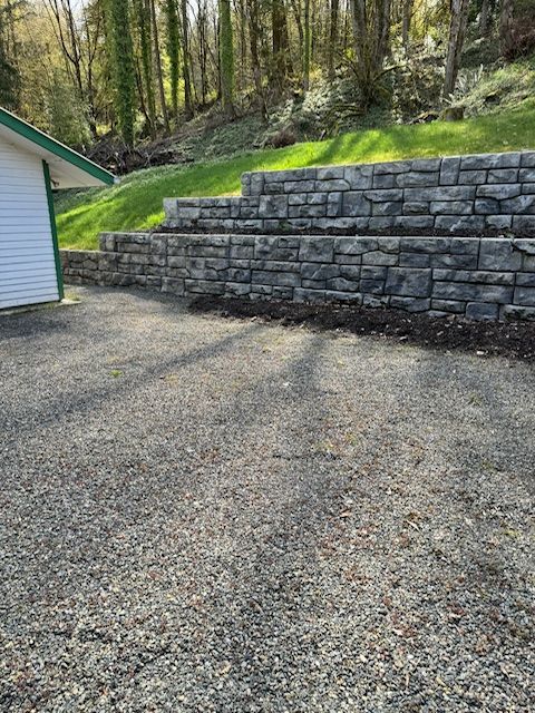 A grey, tiered stone retaining wall stands behind a gravel driveway, bordered by a green hillside and trees.