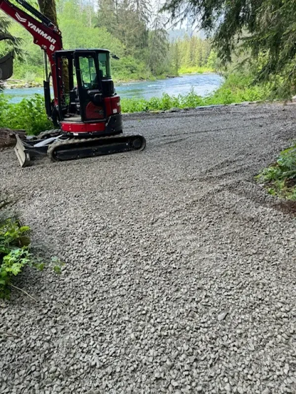 A red Yanmar mini excavator parked on a gravel clearing near a river surrounded by trees.