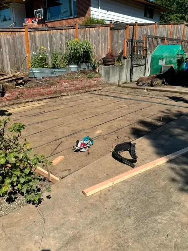 A construction site in a backyard with a circular saw, tool belt, and lumber on a dirt area near a fence and garden bed.
