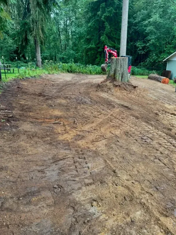 A construction vehicle works on a cleared, muddy lot with tire tracks, surrounded by trees and a building in the distance.