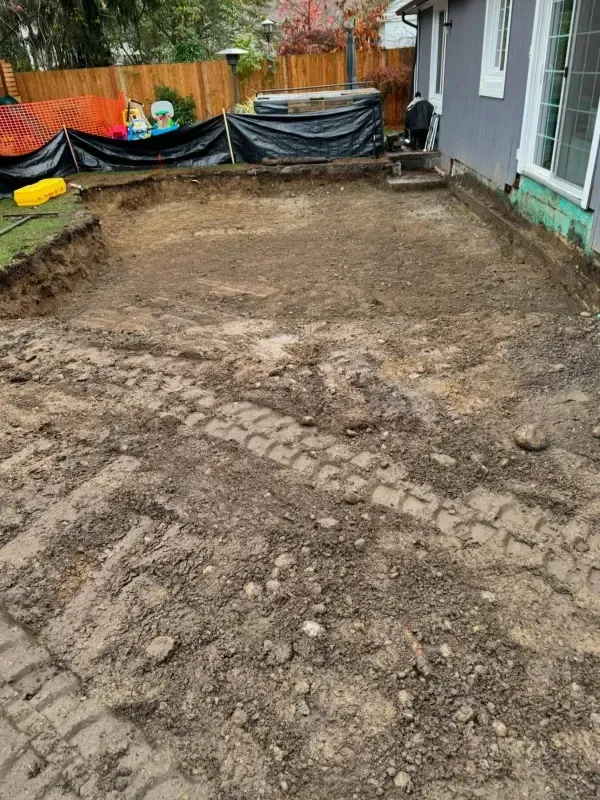 An excavation site in a backyard with exposed dirt, tire tracks, and a black tarp along the fence near a house exterior.