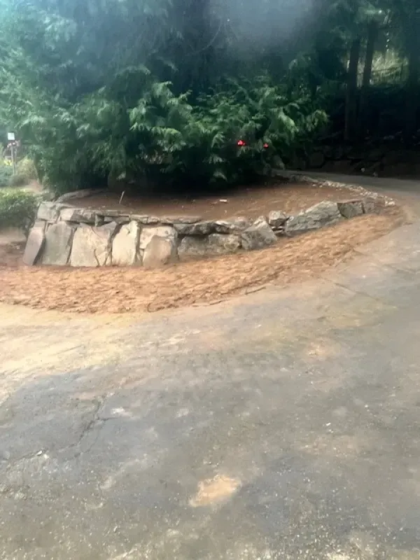 A stone retaining wall surrounds a circular garden bed with green shrubs, situated beside a paved driveway.