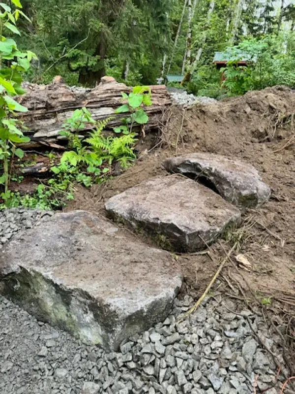 Three large flat stone steps set into a dirt embankment, transitioning from a gravel path into a forested area.