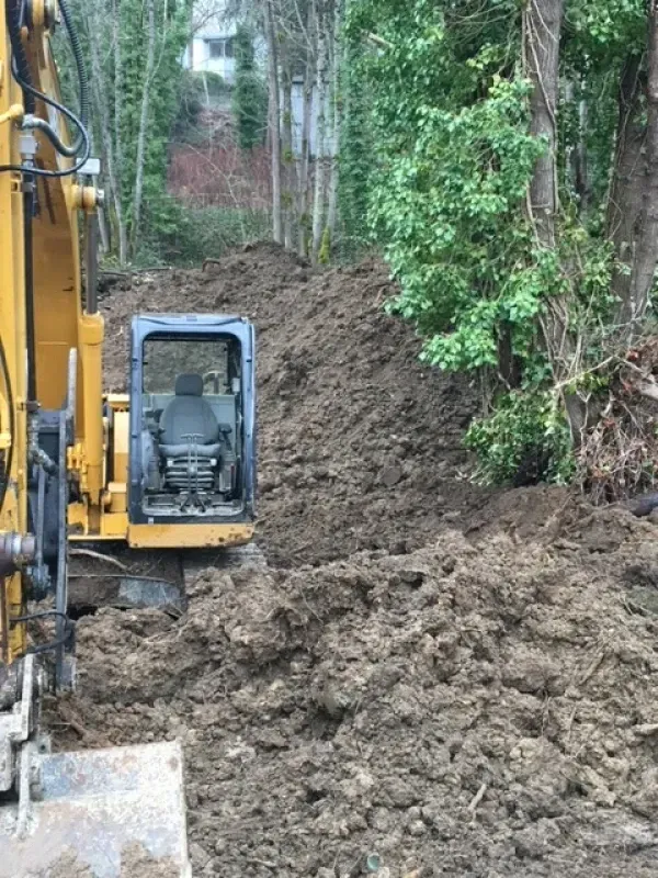 Yellow excavator parked at a construction site with mounds of dirt and trees in the background.