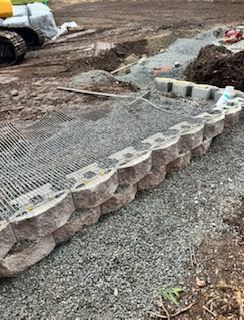 A retaining wall under construction with stacked concrete blocks and gravel on a dirt site near construction equipment.