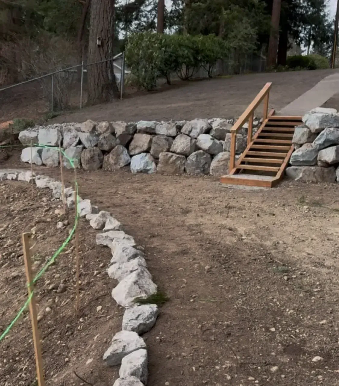 A stone retaining wall with wooden stairs leading up a sloped yard toward a driveway.