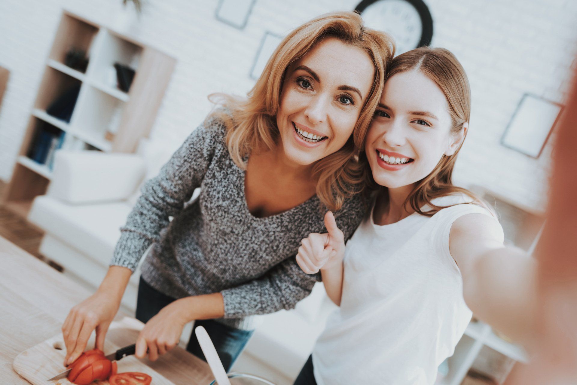 A proud mom and her teen daughter showing their kitchen skills. The mom is cutting fresh tomatoes while the daughter gives a thumbs up, indicating their approval of the meal they're preparing. The two are standing side by side, smiling and happy to be cooking together. This image captures the bond and love between a mother and daughter and their shared love of cooking and creating together.