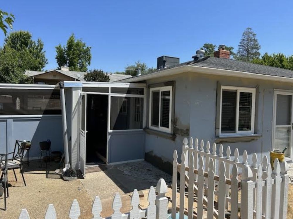 Small backyard patio beside a light gray house, with a white picket fence and outdoor chairs under a canopy.