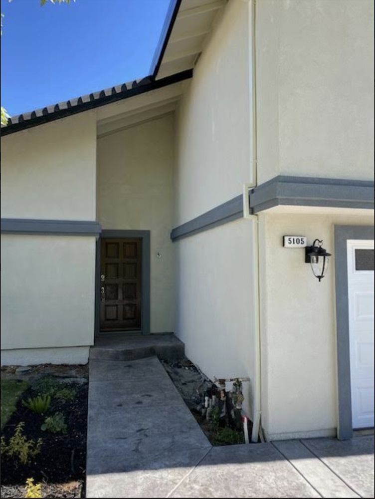 Front entryway of a cream-colored house with a narrow walkway, door, and wall-mounted lantern.