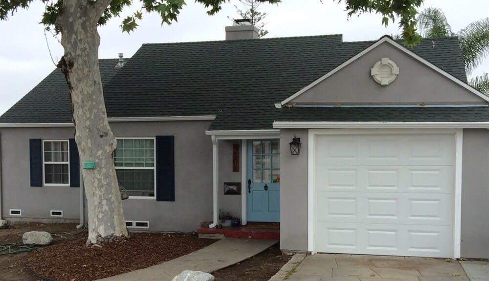 Single-story gray house with a white garage door, blue front door, and a tree in the front yard