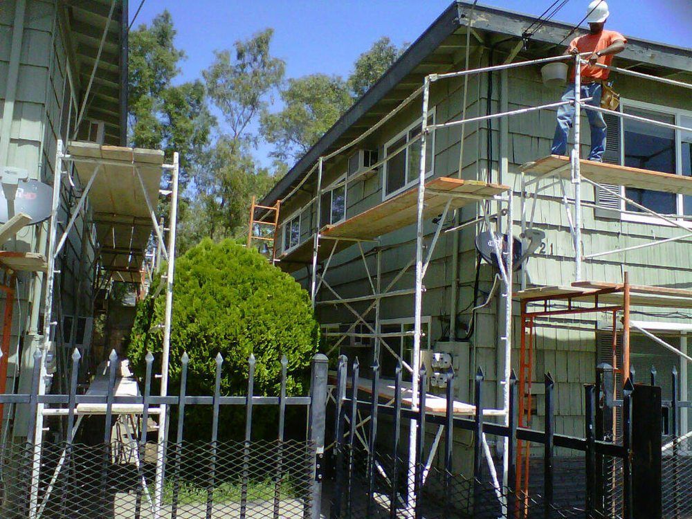 Two-story house under renovation with scaffolding and a worker on the roof, behind a chain-link fence