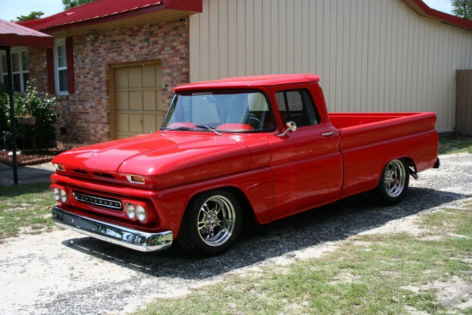 A bright red, vintage Chevrolet pickup truck with chrome wheels parked on a gravel driveway in front of a house.