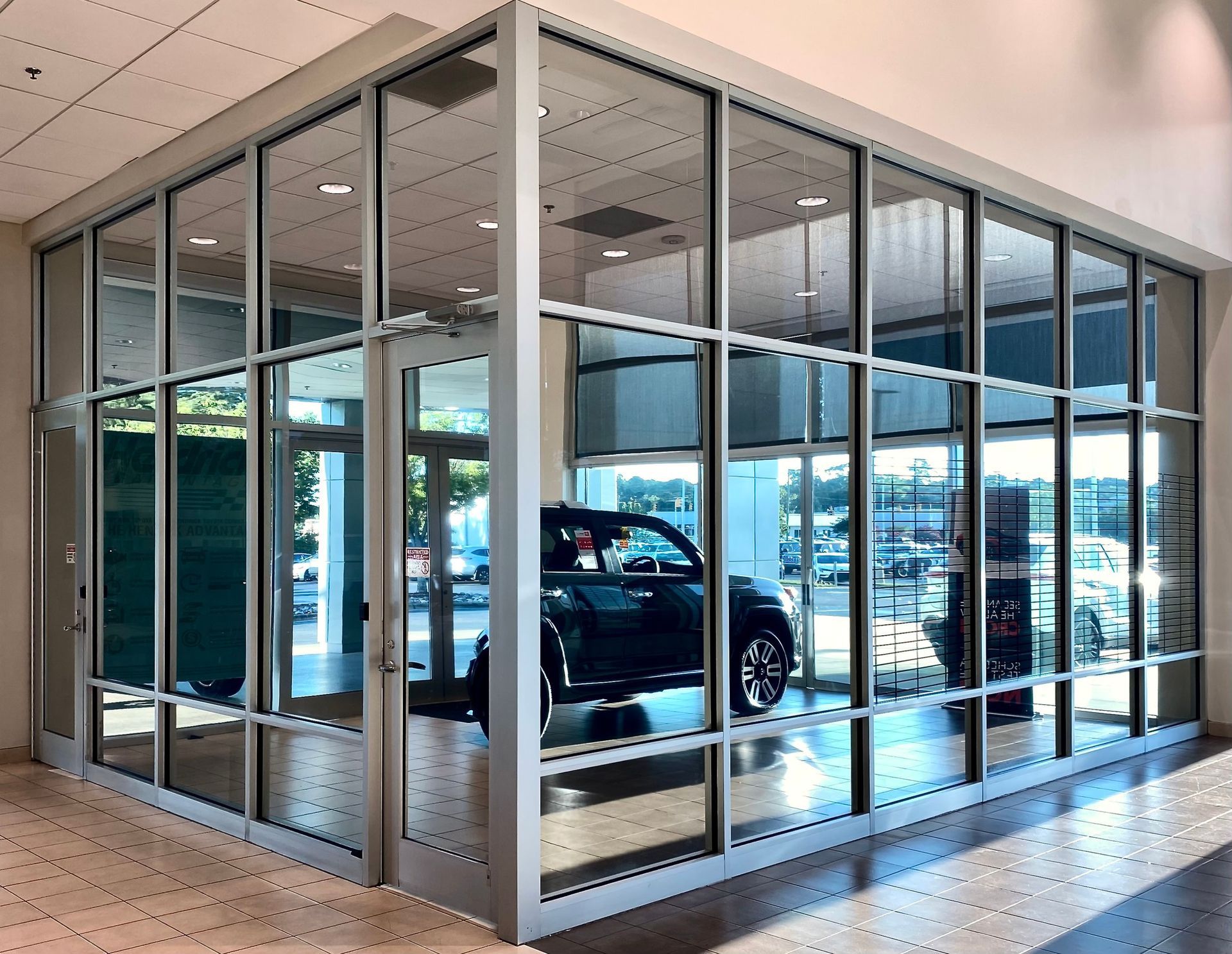A modern glass-walled car dealership display room featuring a dark vehicle indoors, viewed from an adjacent tile hallway.
