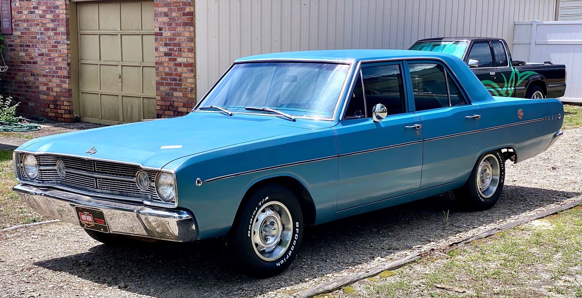 A blue 1960s-era four-door sedan parked on a gravel driveway in front of a brick building.