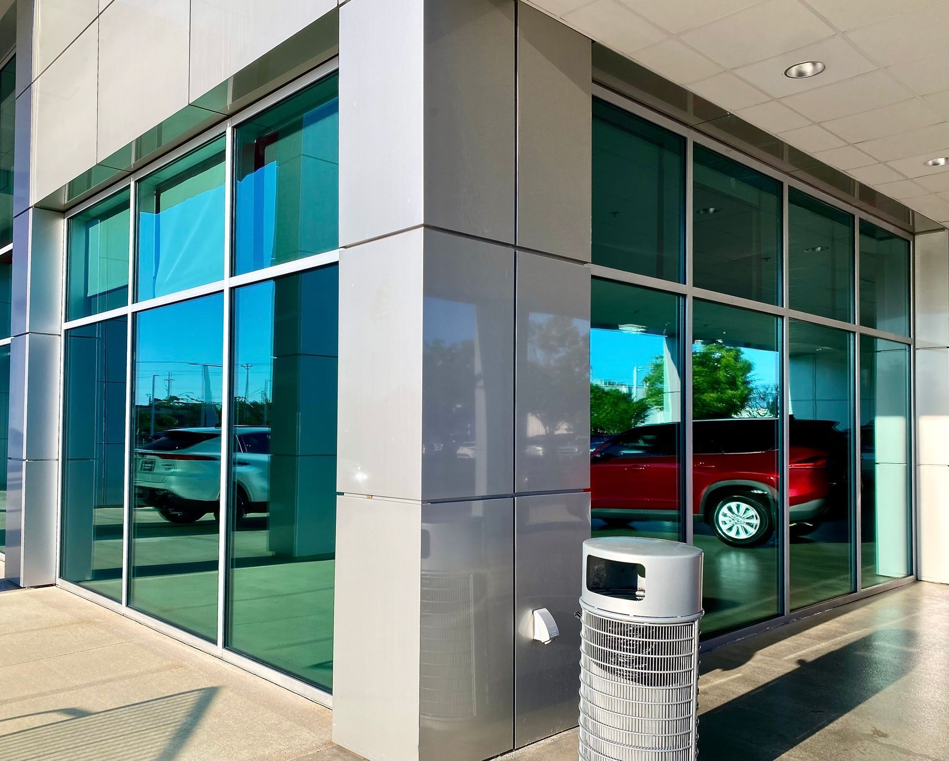 Corner view of a modern glass-walled car dealership showroom featuring a silver exterior, red car, and trash receptacle.