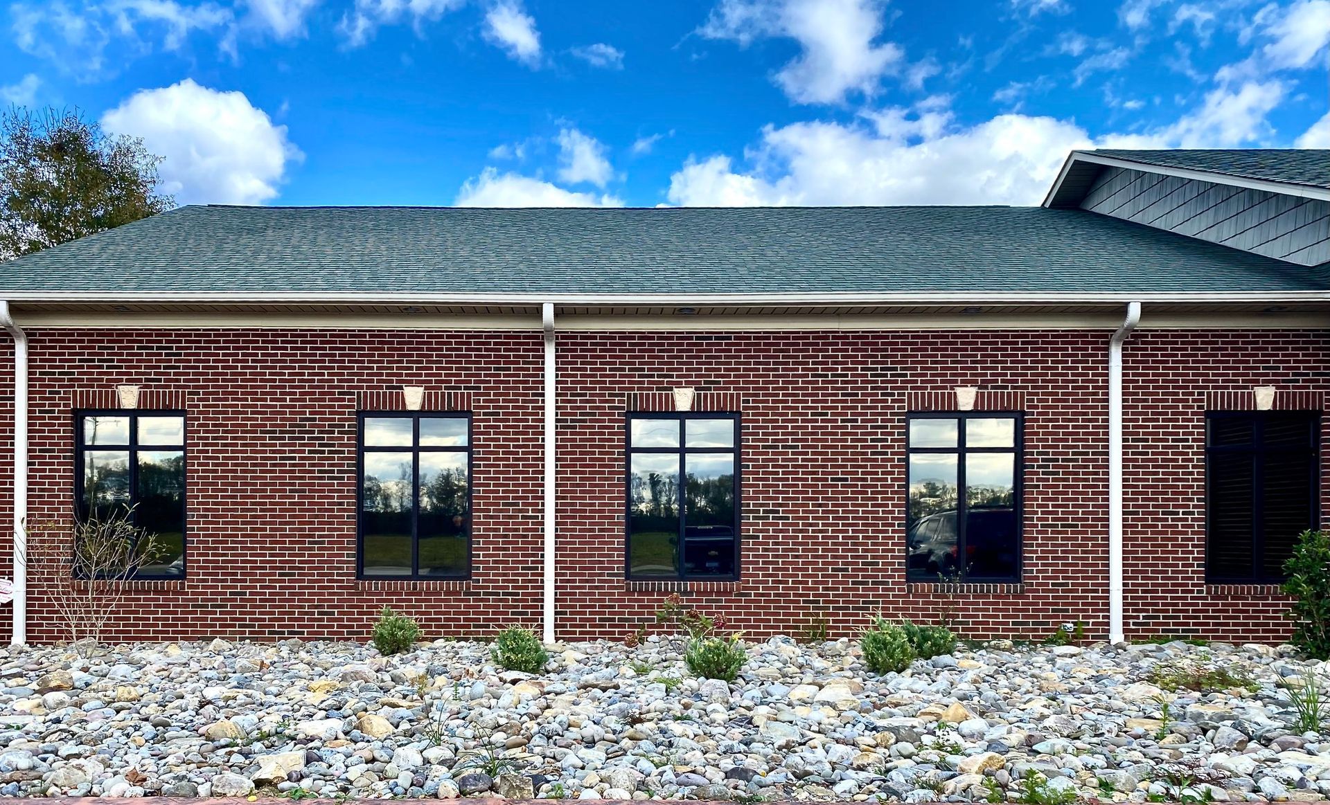 A single-story brick building with five dark-tinted windows, white trim, a dark shingled roof, and a rock-covered lawn.