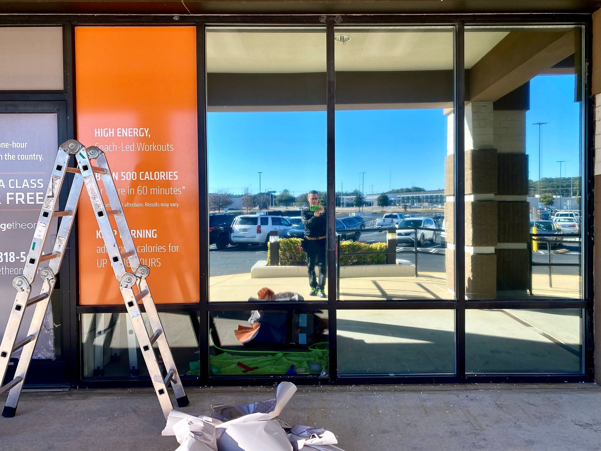 A metal ladder leans against an orange storefront window, with a person reflected in the glass and supplies on the ground.