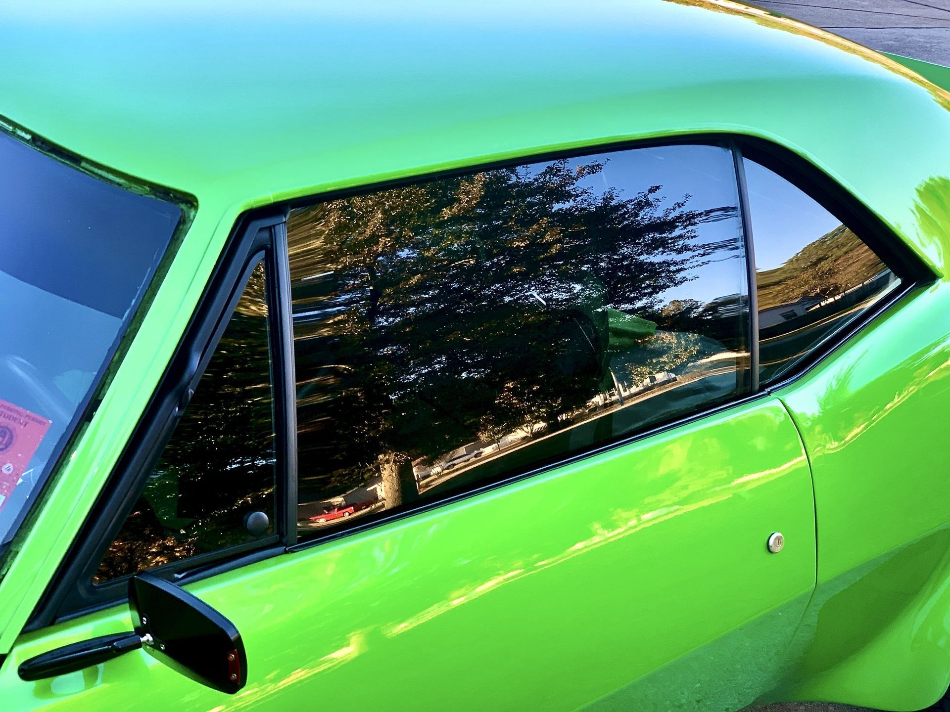 A close-up view of the window and side mirror of a bright green muscle car, reflecting trees and sky.