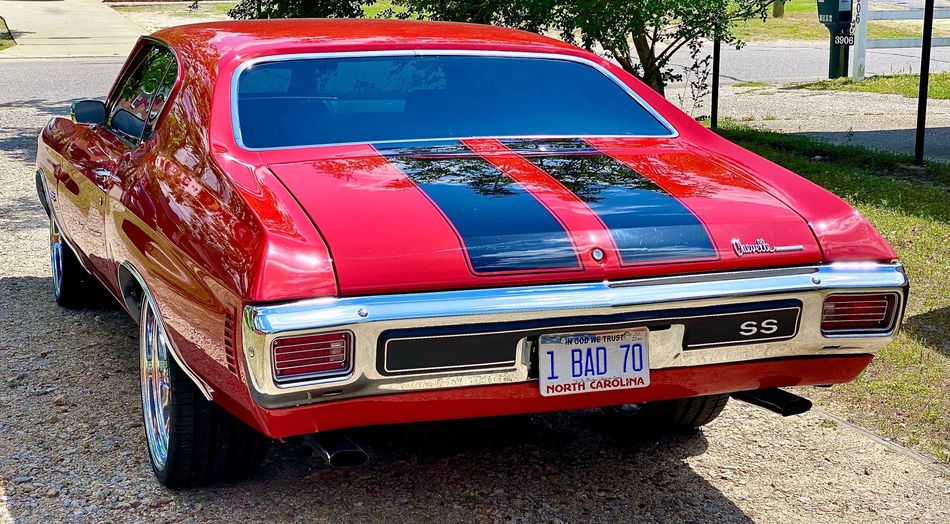 A red Ford Mustang with black racing stripes parked on a gravel drive next to a brick wall and green trees.