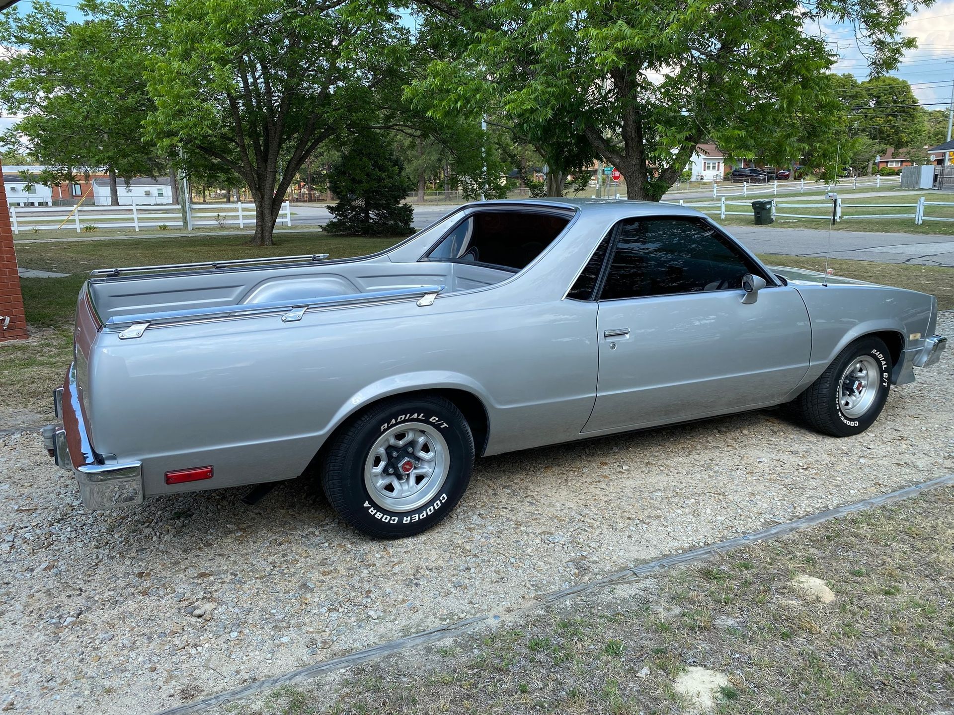 A silver Chevrolet El Camino parked on a gravel surface outdoors with trees in the background.