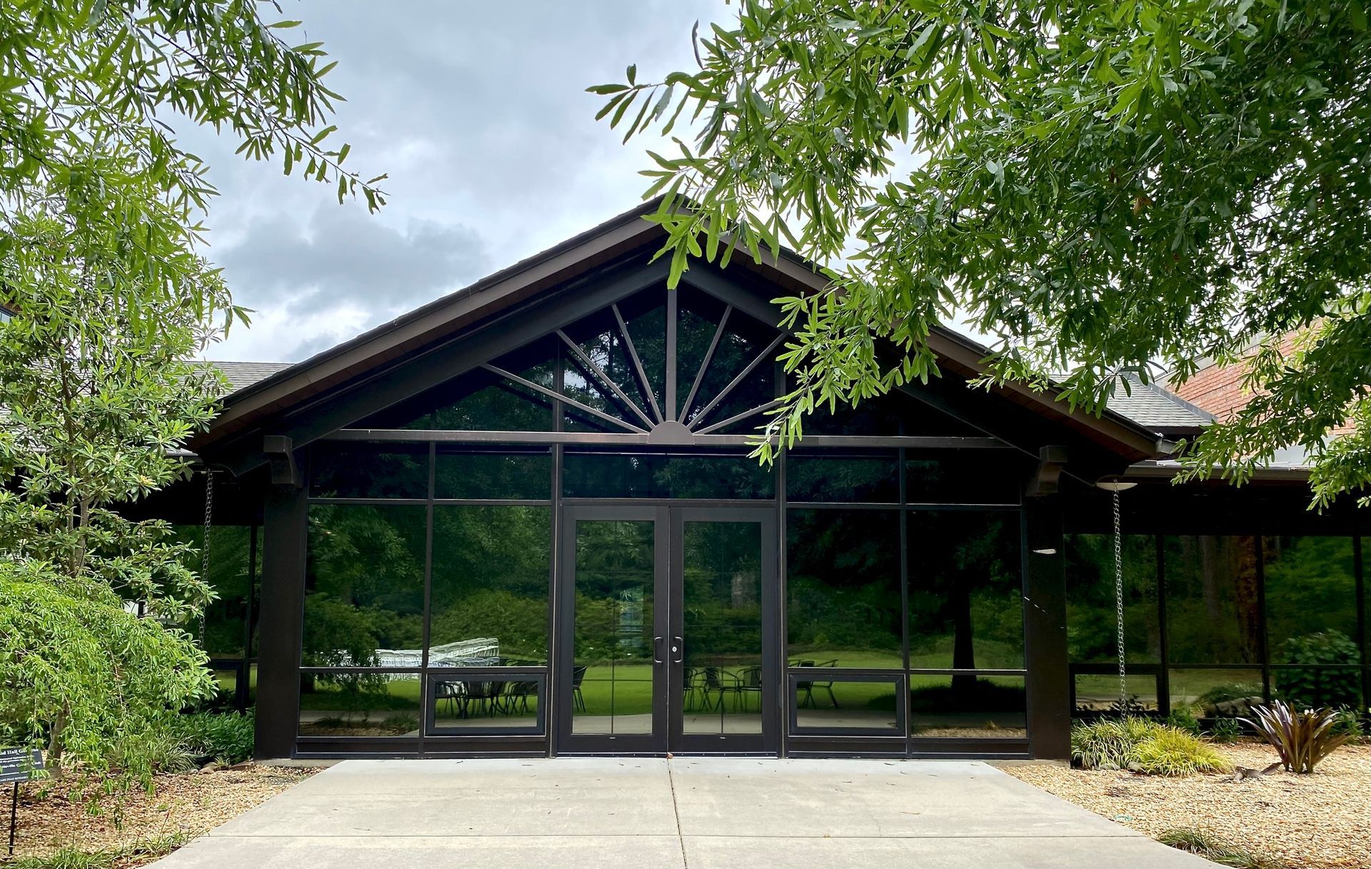 A dark-framed building entrance with floor-to-ceiling windows and a sunburst-style gable window surrounded by lush trees.
