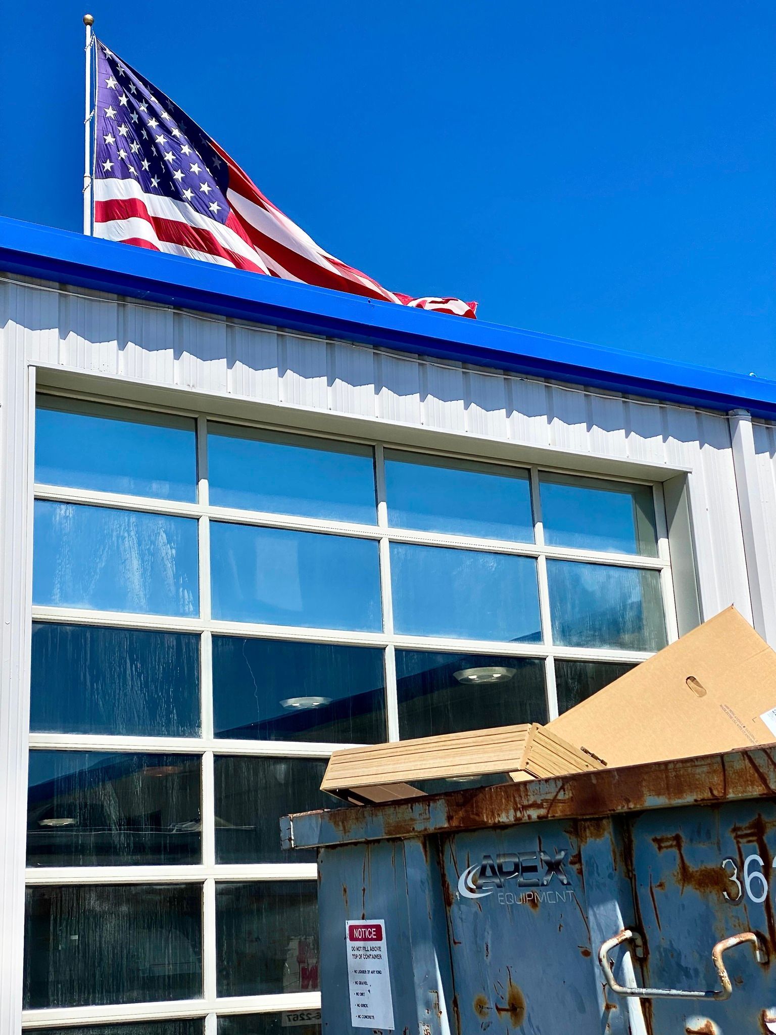 A blue dumpster sits in front of a glass-paneled garage door with an American flag flying on the roof against a blue sky.