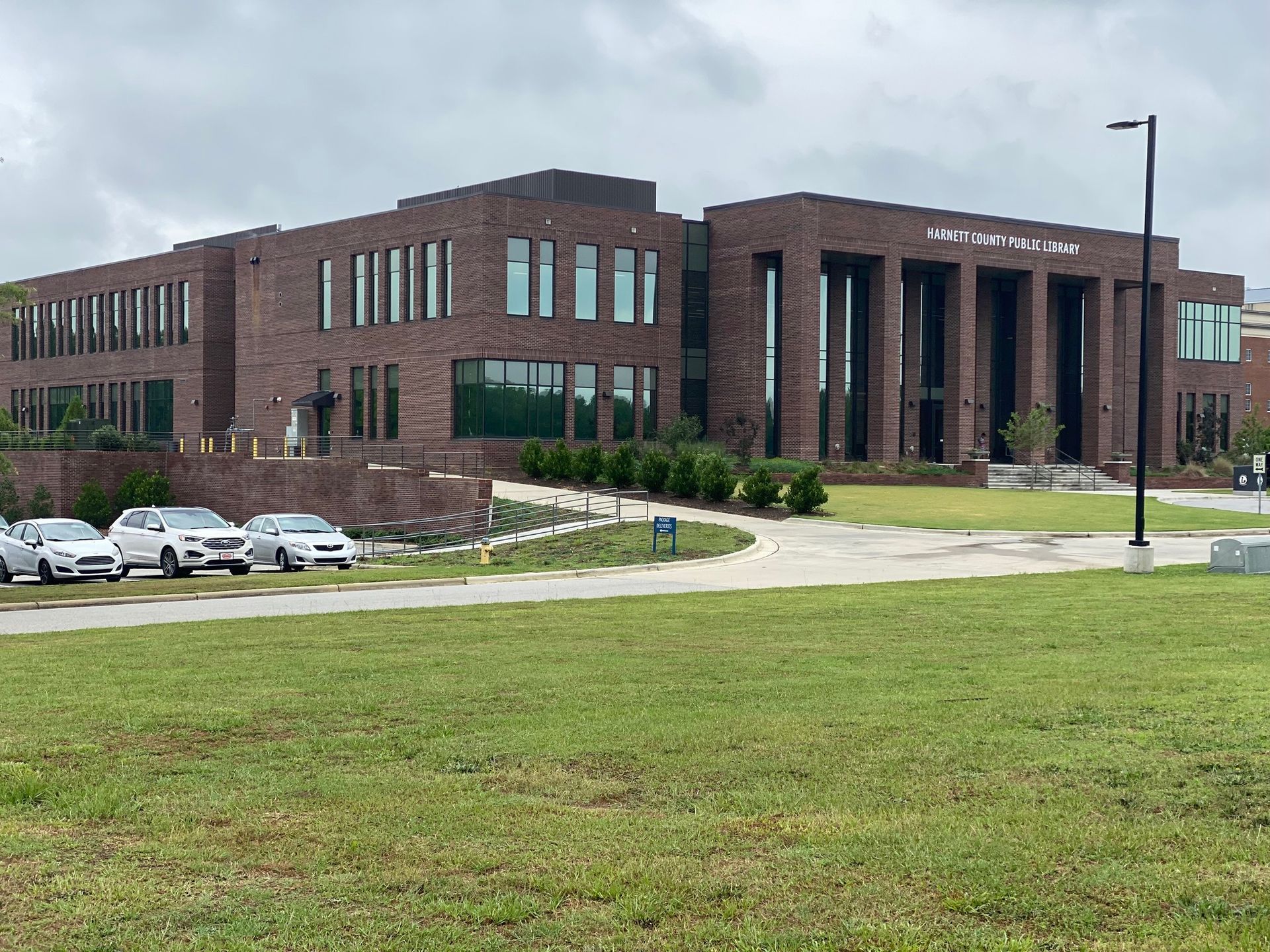 A two-story dark brick building with a columned entrance and modern windows, set behind a lawn and a parking area.