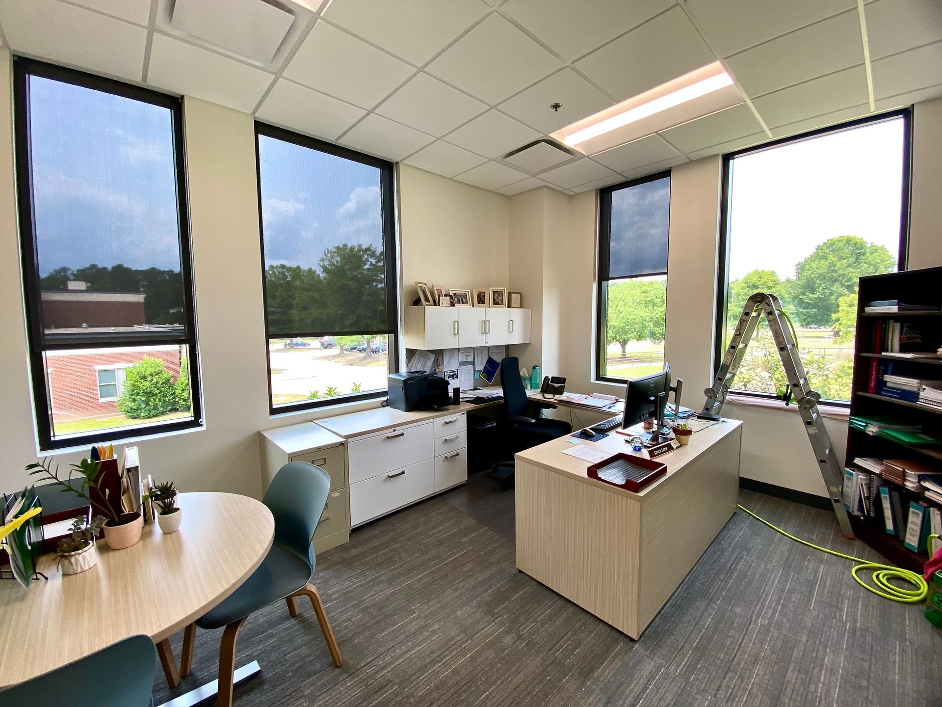A bright office with desks, chairs, a bookshelf, and a ladder leaning against a window with a sunshade.