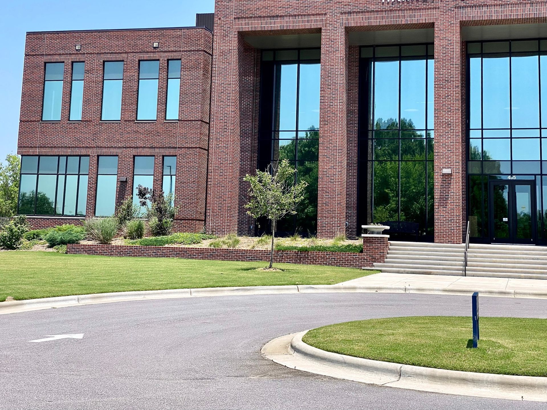 A multi-story building featuring dark red brick, tall glass windows, and a paved entrance area with a grassy foreground.