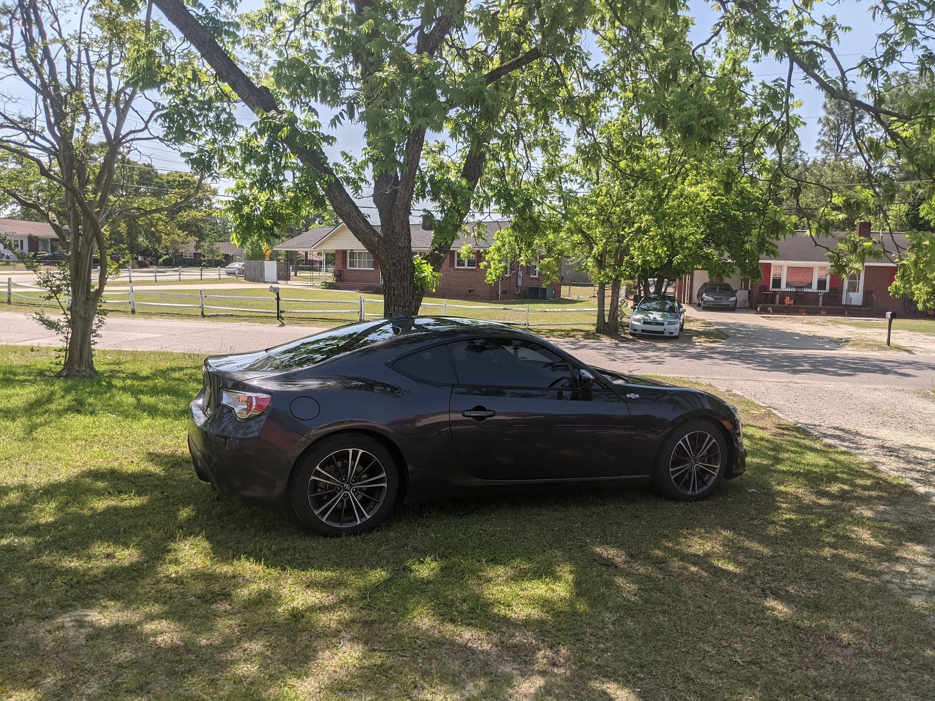 A dark gray two-door sports car parked on the grass beneath a large tree in front of a residential house.