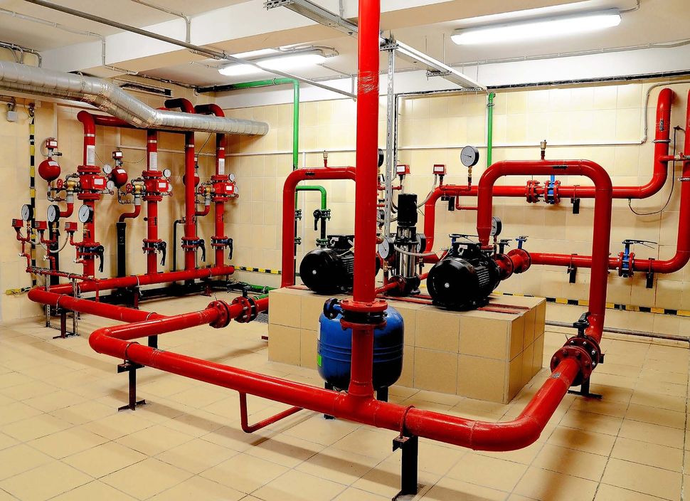 Red industrial pipes and valves in a mechanical room with pumps and beige tile floor