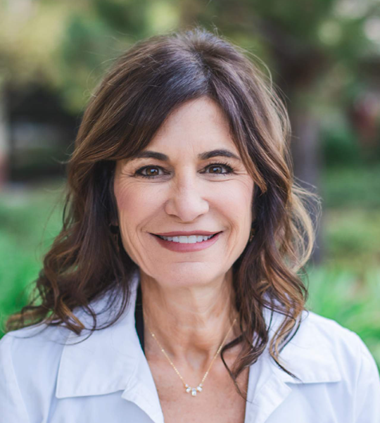 A smiling woman with long brown hair, wearing a white shirt and a delicate necklace, standing against a blurred backdrop.