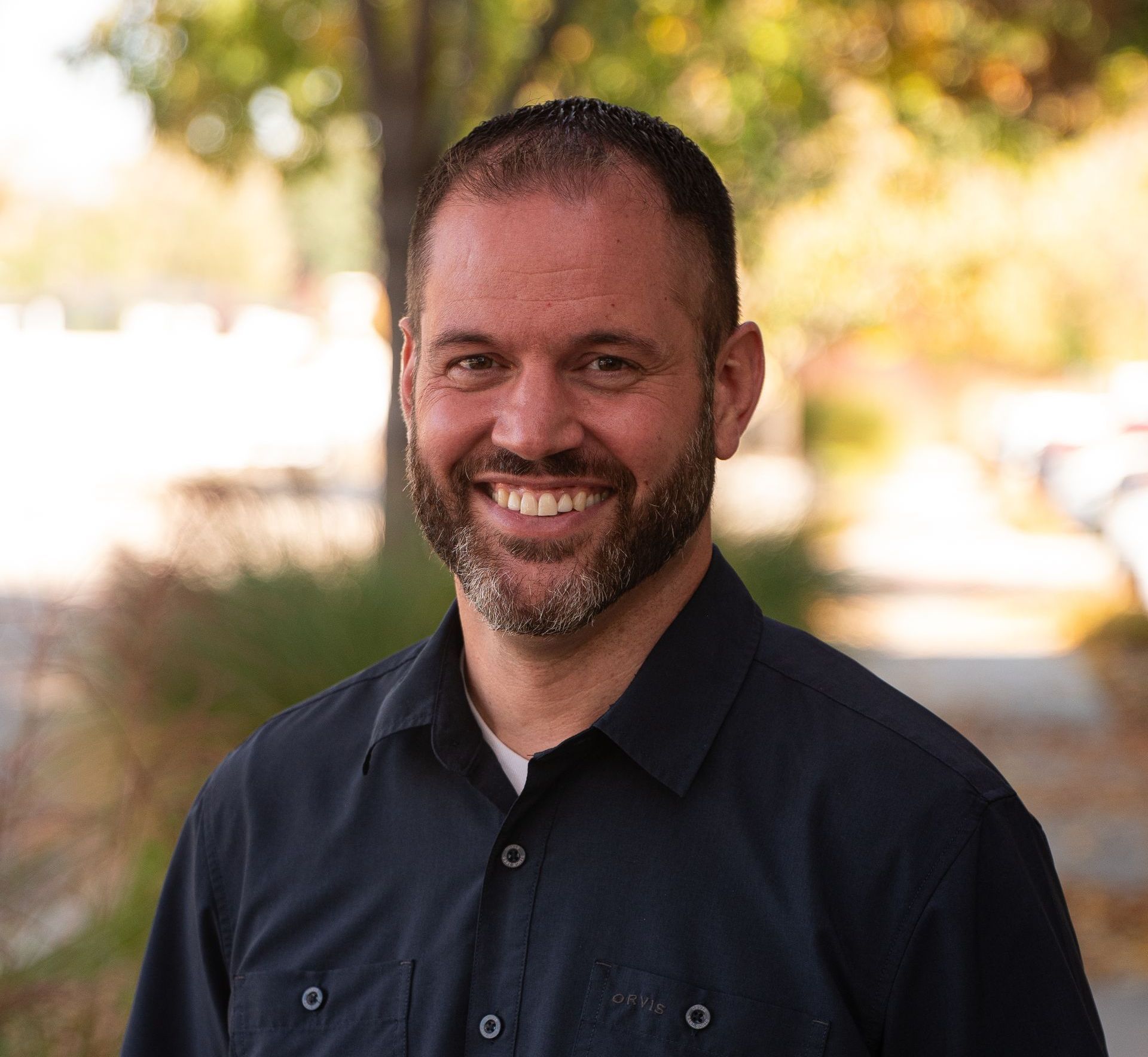 A smiling person with a beard, wearing a dark button-down shirt, standing outdoors with a blurred background.