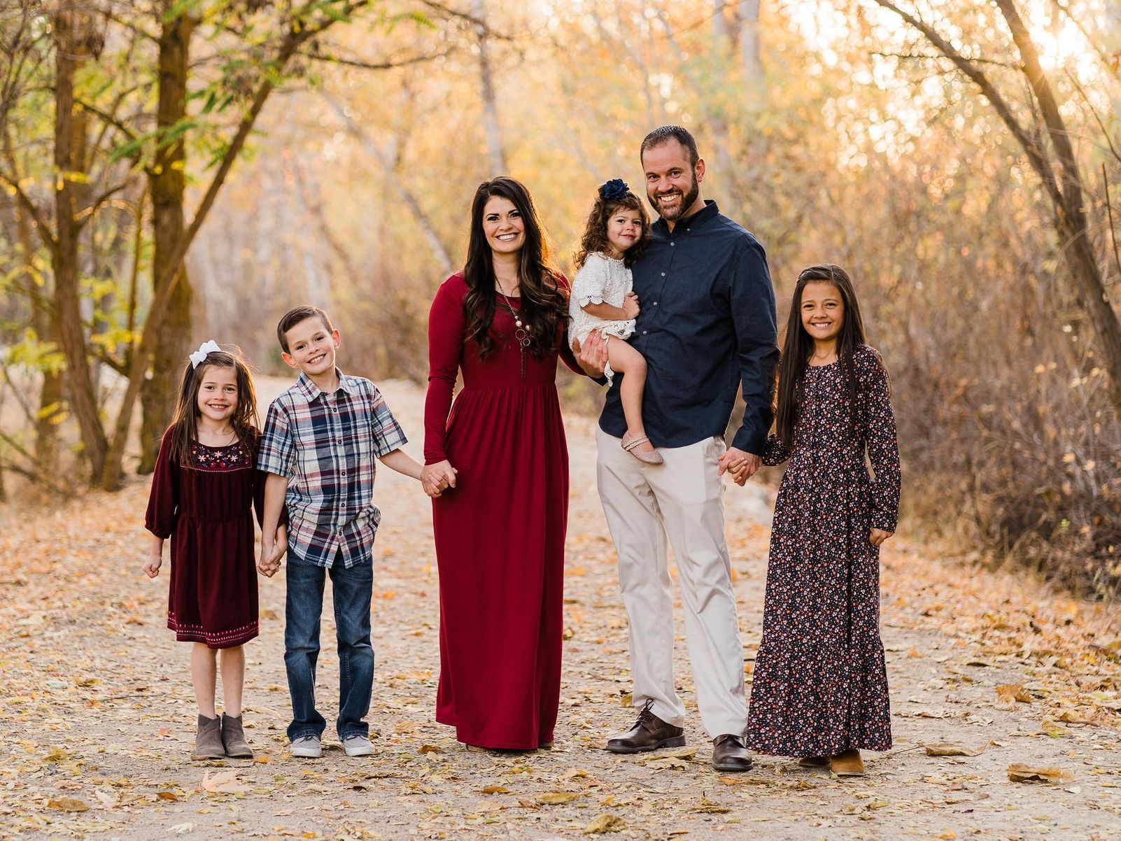 A family of six smiling while standing together on a sunlit, leaf-covered path in an autumn forest.