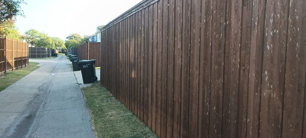 Narrow sidewalk beside a long wooden fence, with trash bins and trees in the distance.
