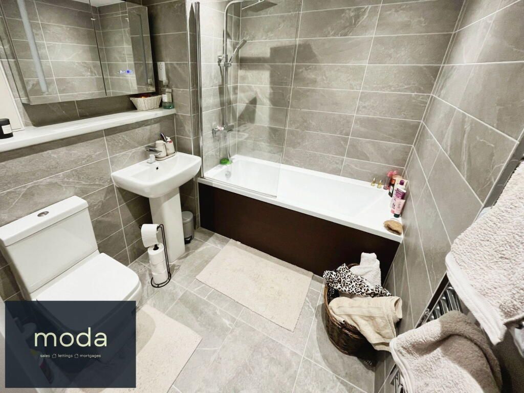 Bathroom with gray tiled walls, white toilet, sink, and tub. Brown tub front. Two bathmats and towels.
