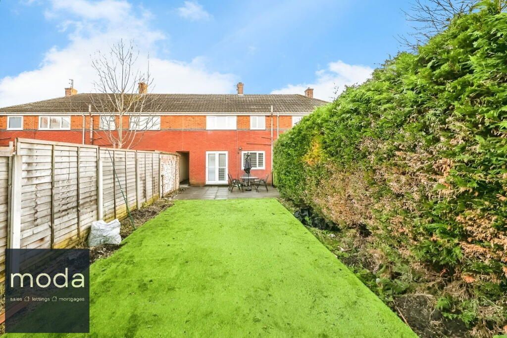 Backyard of a red brick home with green grass, tall hedges and a wooden fence.