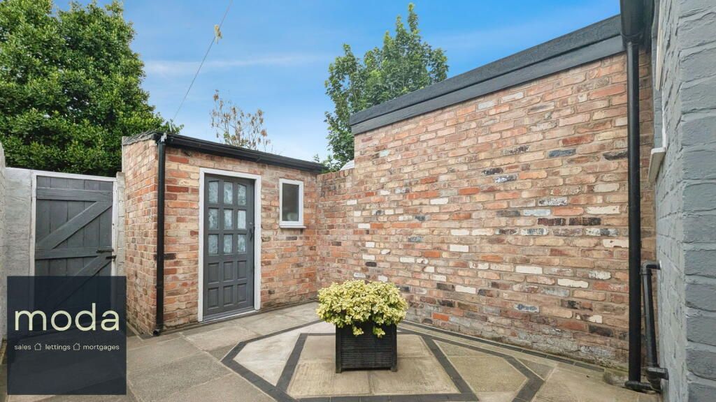 Exterior view of a brick-walled patio with a shed, a pot of plants, and a gray door under a blue sky.