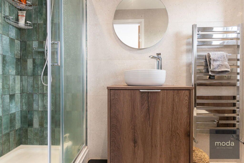 Bathroom with a wood-tone vanity, white sink, round mirror, heated towel rack, and green tiled shower.