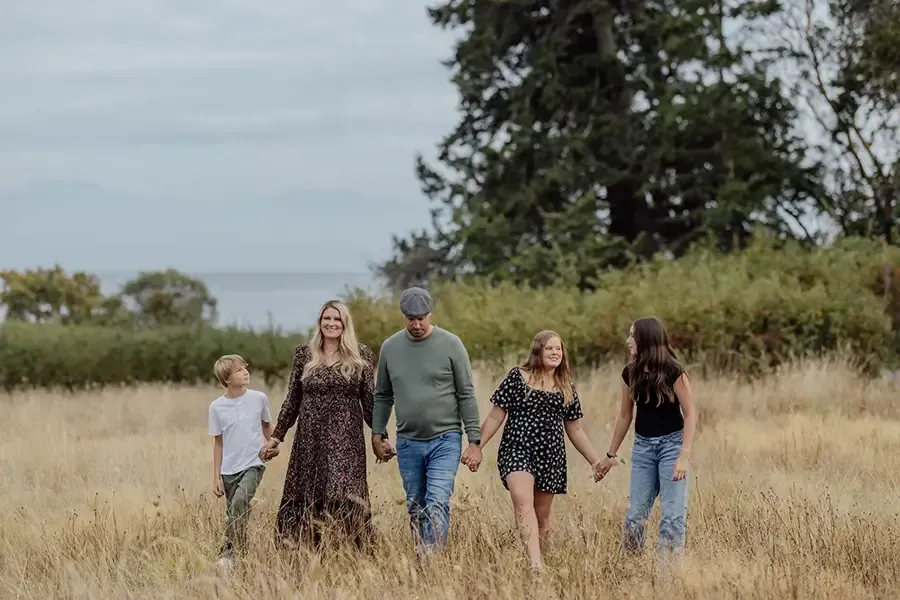 Five people walking through tall grass under a cloudy sky, with trees and water in the distance