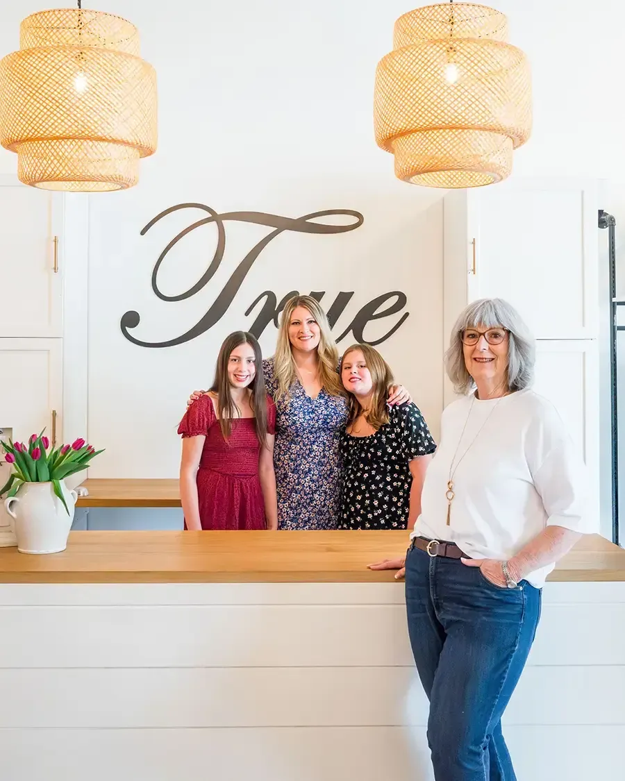 Four women posing in a bright shop with a large “True” sign behind them.