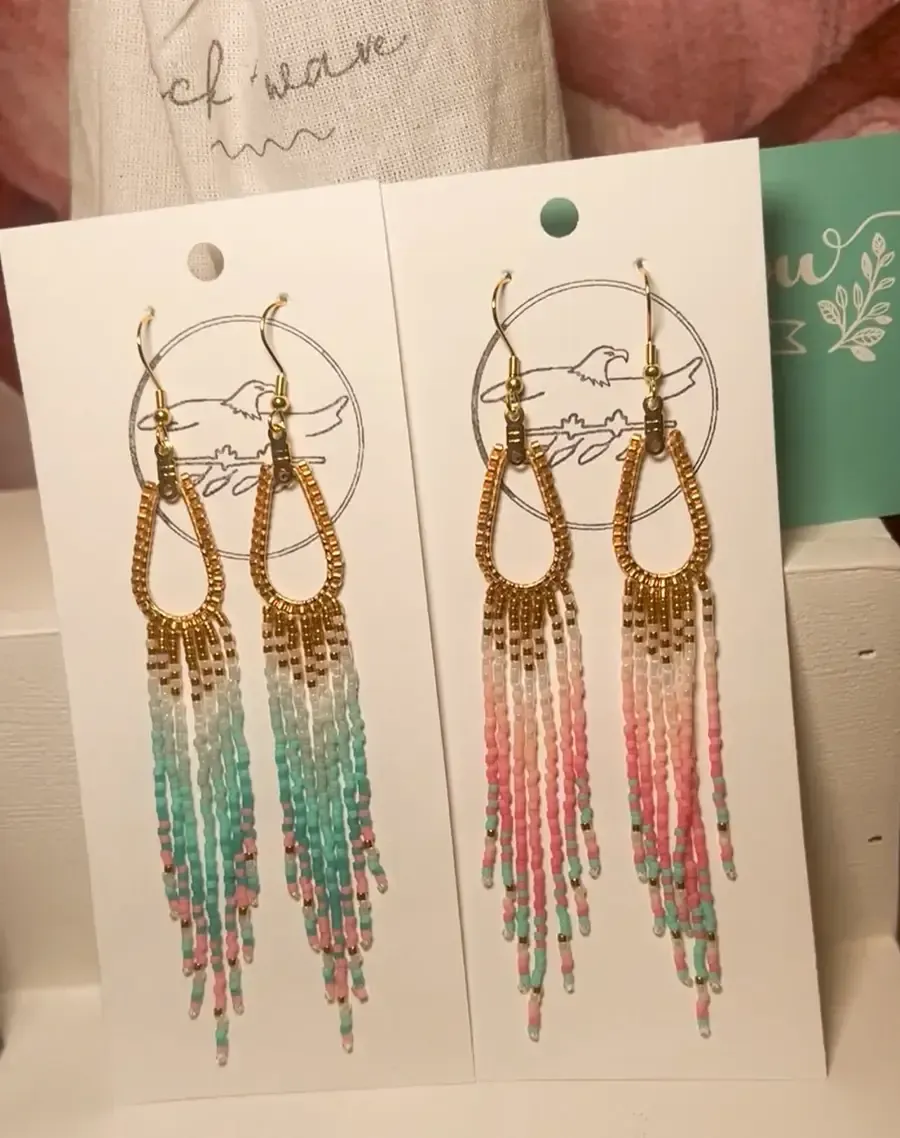 Two pairs of long beaded hoop earrings on white display cards, one teal and one pink, with gold accents