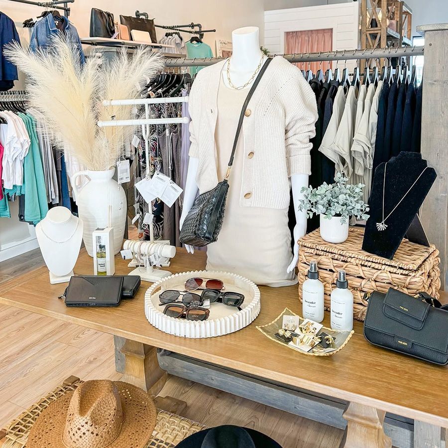 Boutique display with cream sweater outfit, black accessories, woven baskets, and home decor on a wooden table