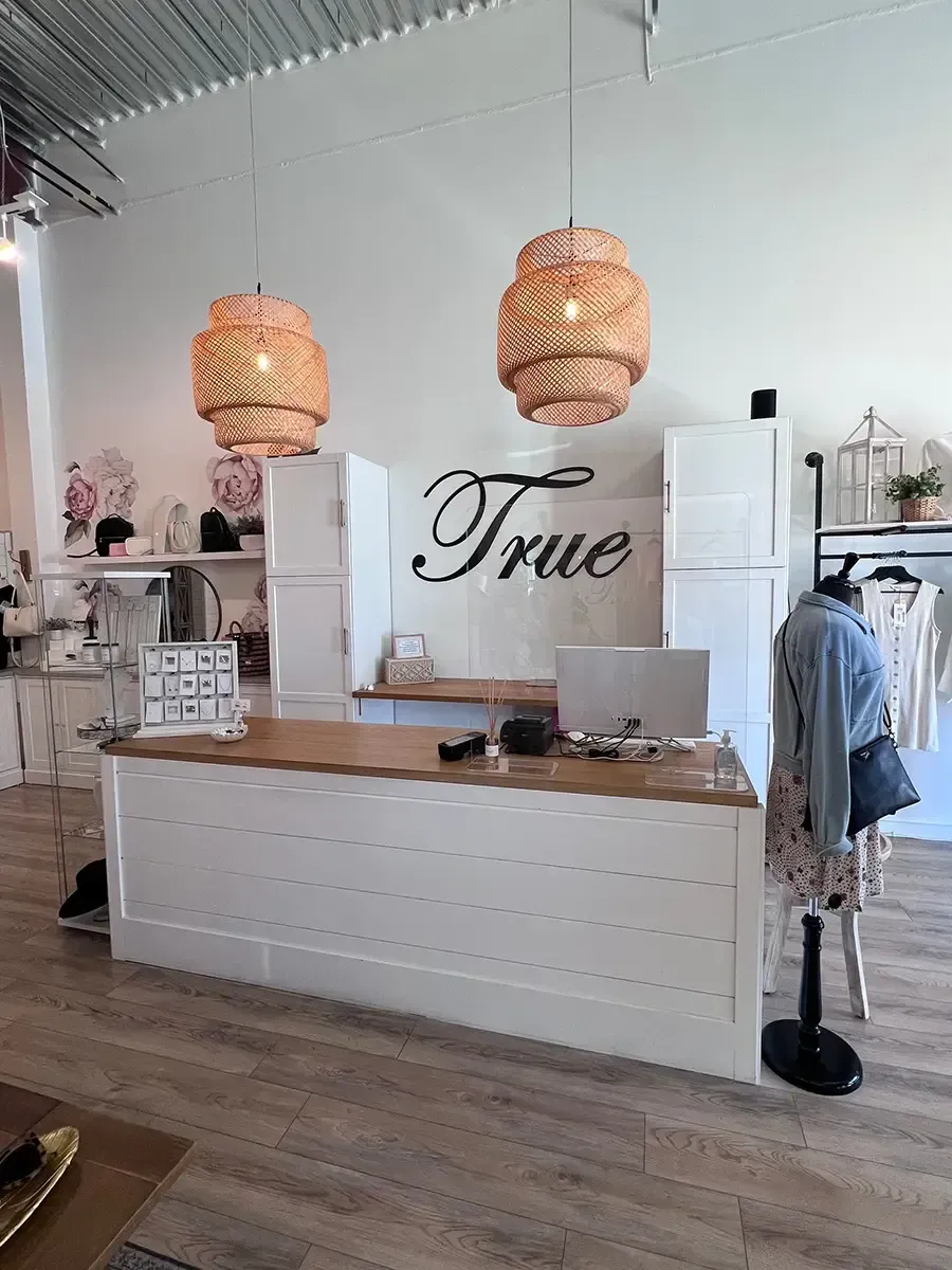 Boutique clothing shop interior with white counter, wicker pendant lights, and “True” sign on the back wall