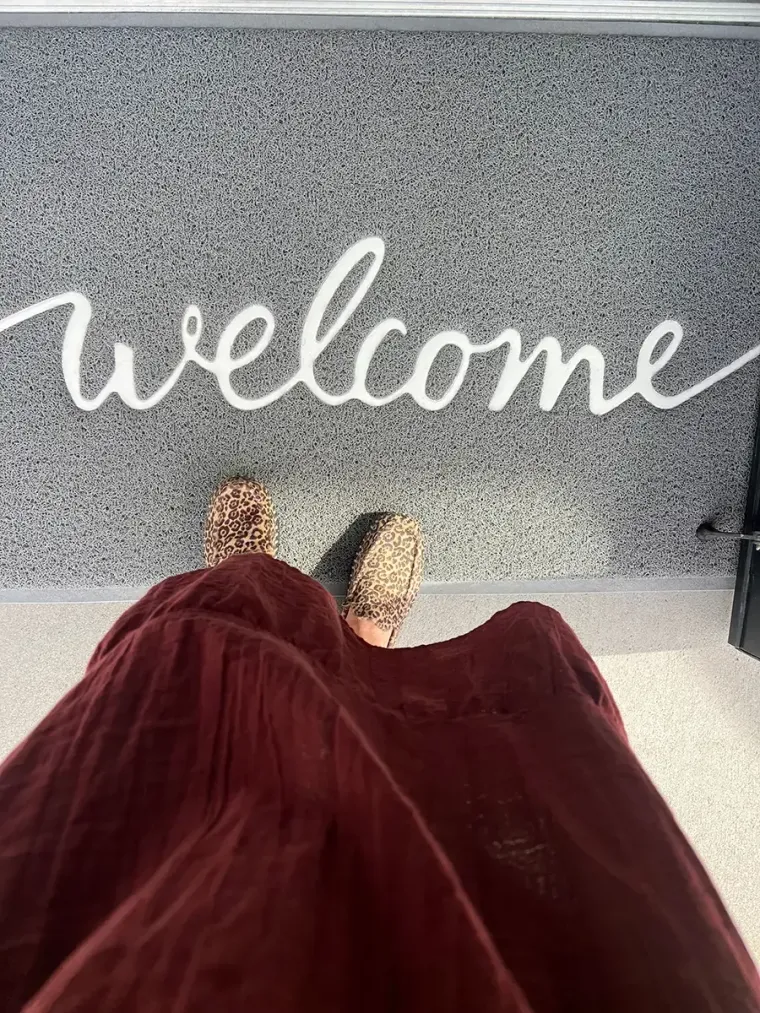 Feet in gold shoes and burgundy skirt facing a gray welcome mat with white script.