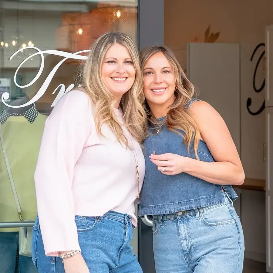 Two smiling women in light blue jeans and casual tops standing in a shop doorway