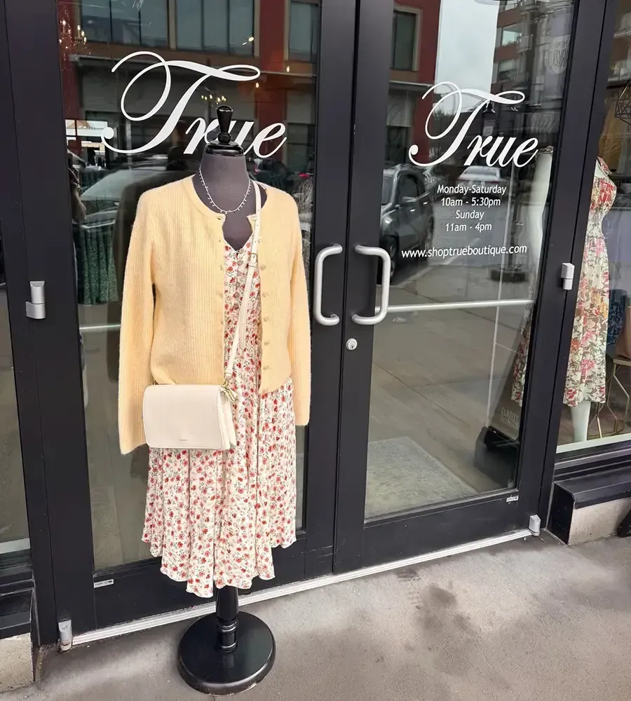 Storefront display with a yellow cardigan, floral dress, and white clutch on a mannequin.